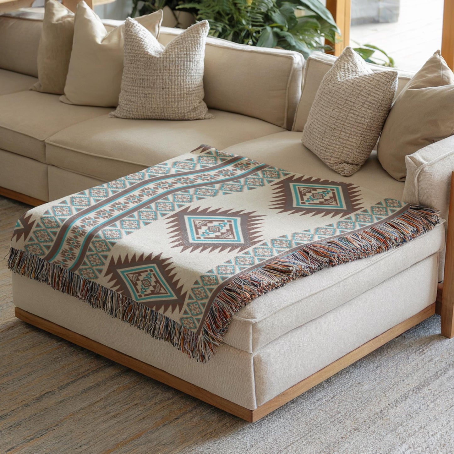 a cozy living room with a beige sofa and a decorative throw blanket with a geometric pattern on it. The sofa is positioned against a wall with a window, and there are several pillows on the sofa. The room also features a rug on the floor and a potted plant in the background.