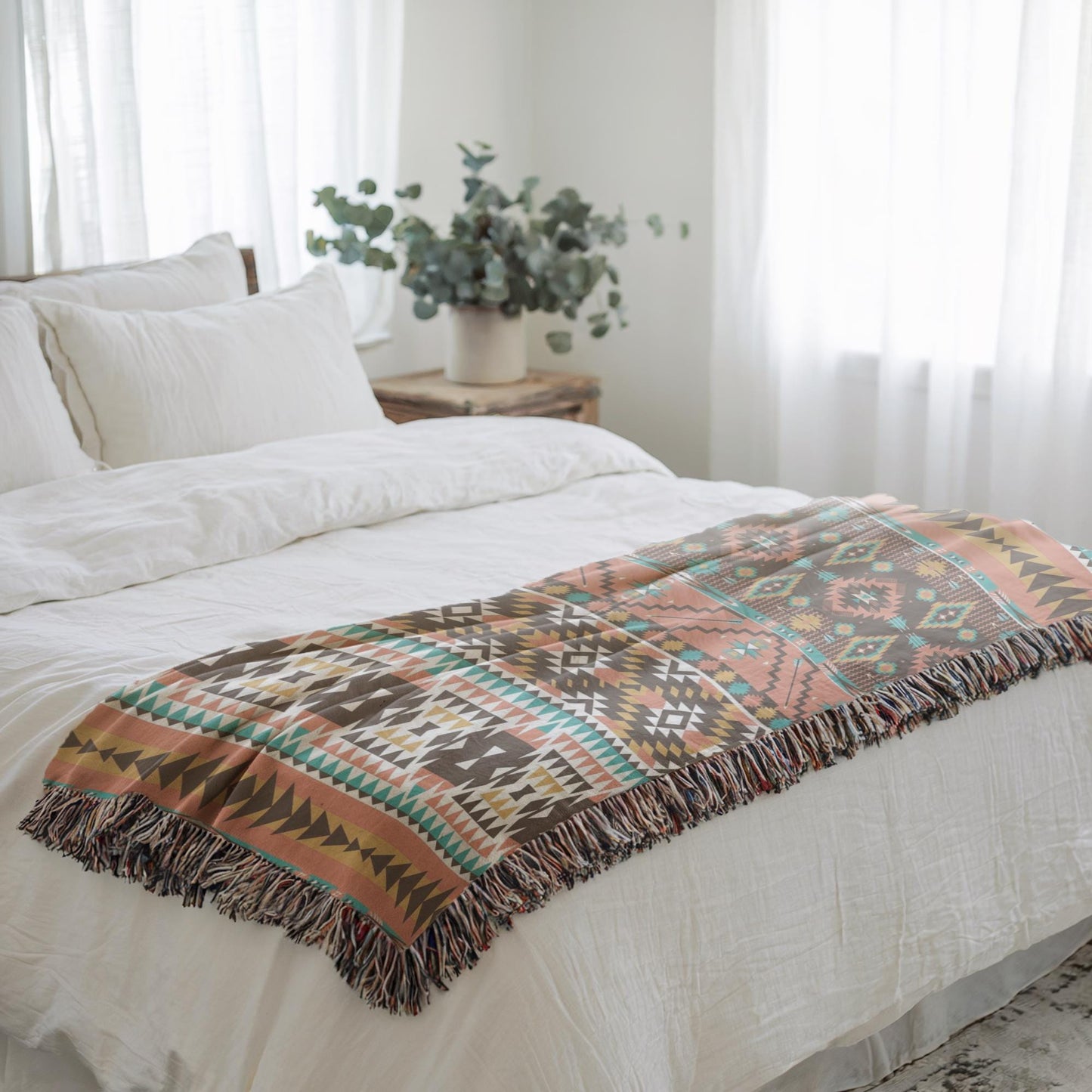 a cozy bedroom scene with a bed covered in a colorful patterned blanket and a white comforter. A wooden nightstand is positioned next to the bed, and a potted plant is placed on the nightstand. The room has white curtains and a window, allowing natural light to fill the space.