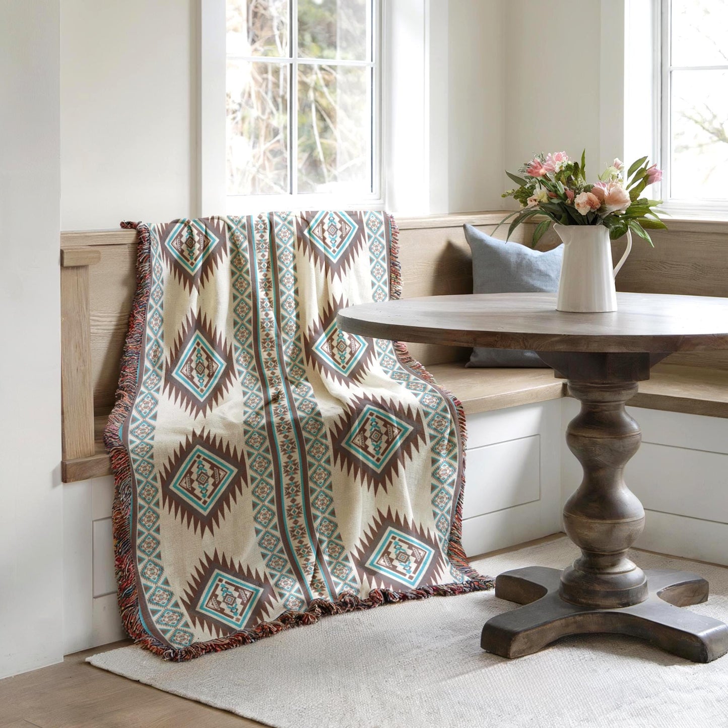 a cozy corner with a wooden table, a bench, and a colorful throw blanket draped over the bench. A vase of flowers is placed on the table, adding a touch of nature to the space. The room has a clean, minimalist aesthetic with white walls and a wooden floor.
