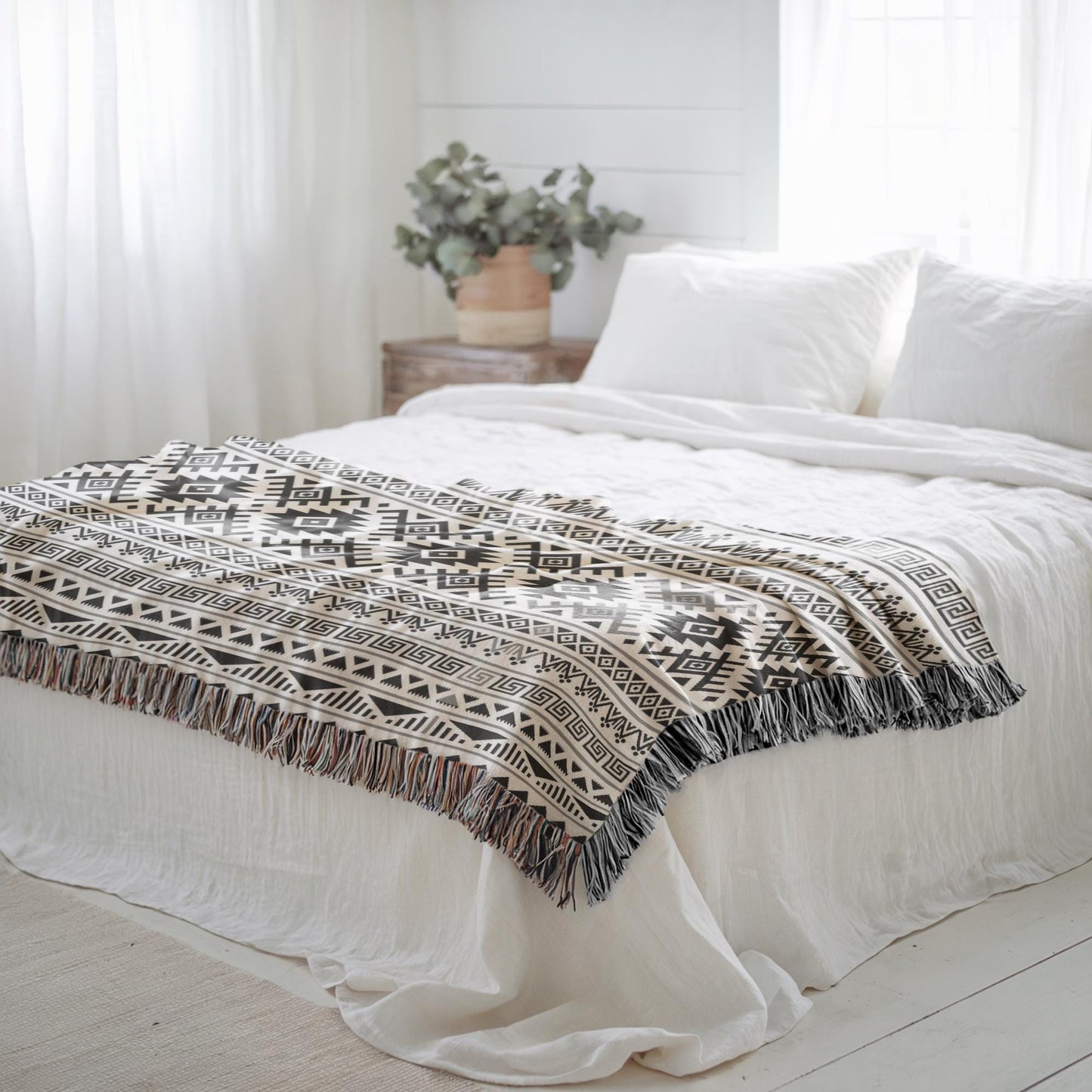 a cozy bedroom with a bed covered in a patterned blanket and white pillows. A potted plant is placed on a nightstand next to the bed, and the room has white curtains and a wooden floor.