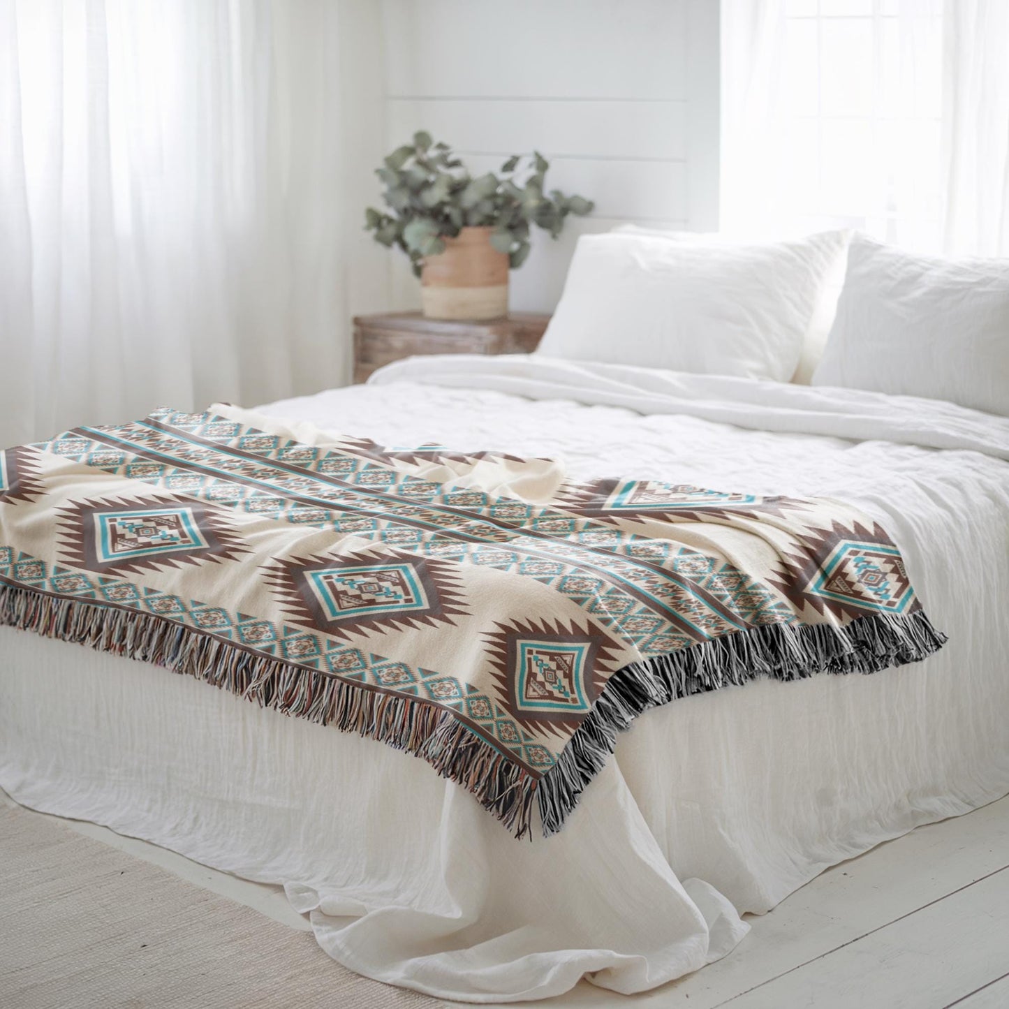a cozy bedroom with a bed covered in a patterned blanket and white pillows. A potted plant is placed on a nightstand next to the bed, and the room has white curtains and a wooden floor.