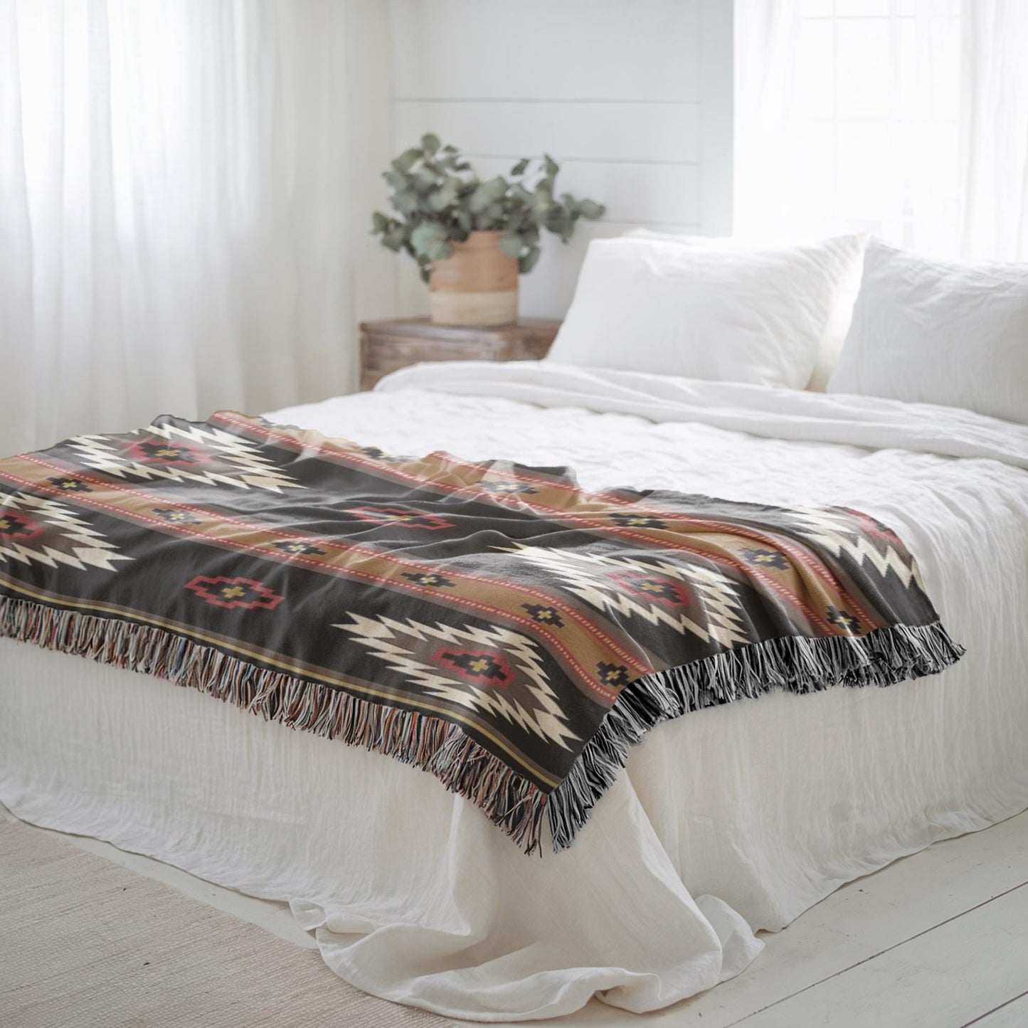 a cozy bedroom with a bed covered in a patterned blanket and white pillows. A potted plant is placed on a nightstand next to the bed, and the room has white curtains and a wooden floor.