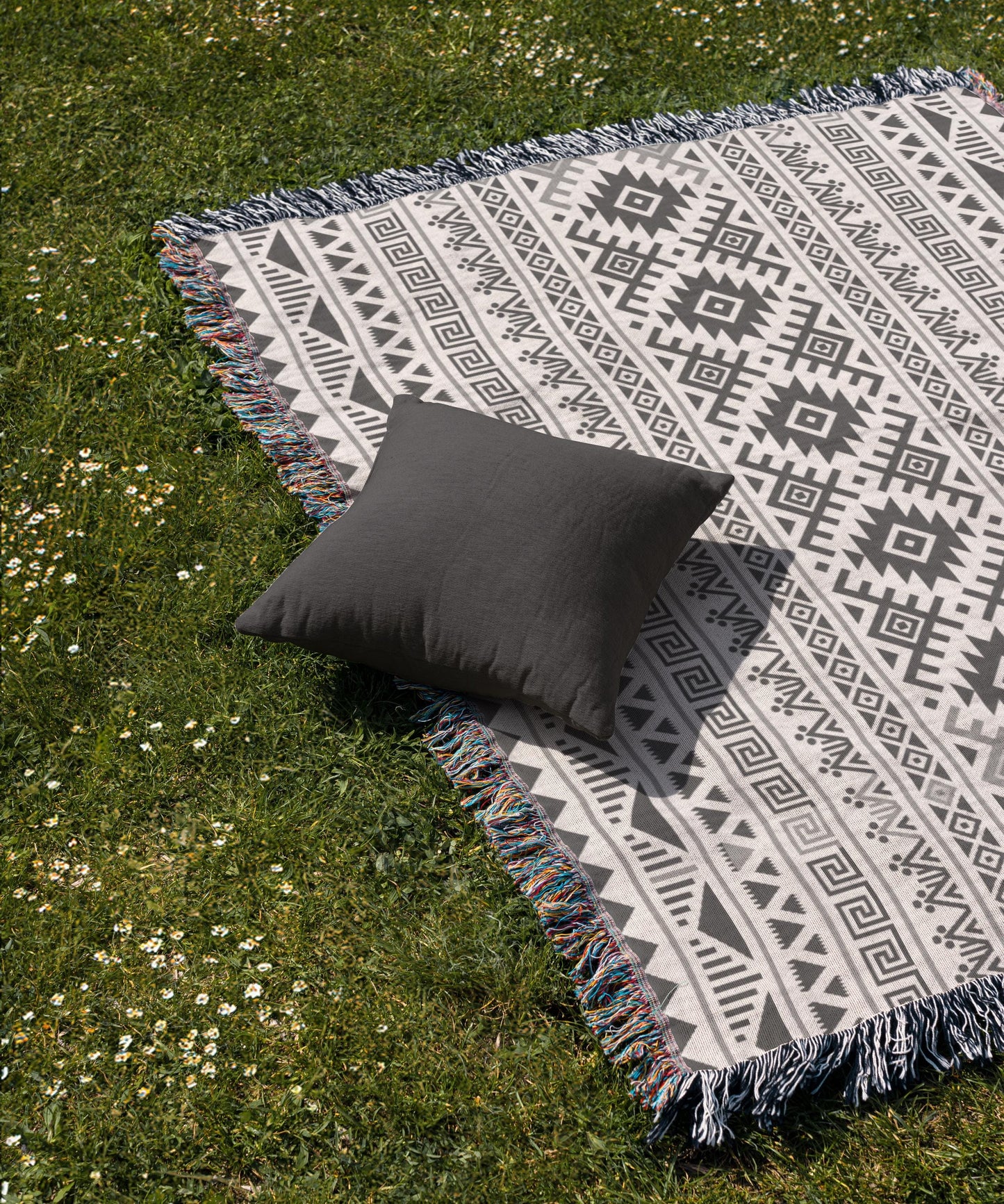 A gray throw pillow rests on a patterned blanket spread out on a grassy lawn.