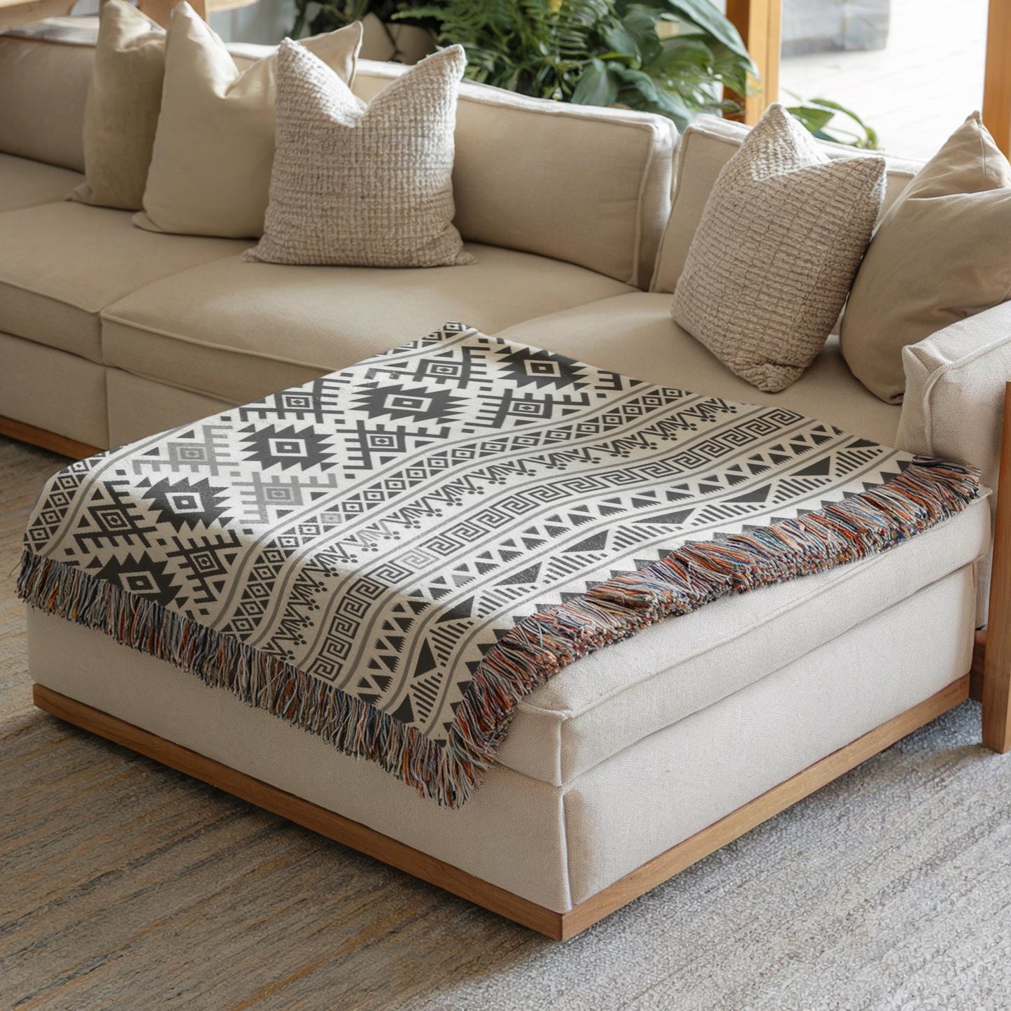 a cozy living room with a beige sofa and a decorative throw blanket with a geometric pattern on it. The sofa is positioned against a wall with a window, and there are several pillows on the sofa. The room has a neutral color scheme with a touch of patterned textiles.