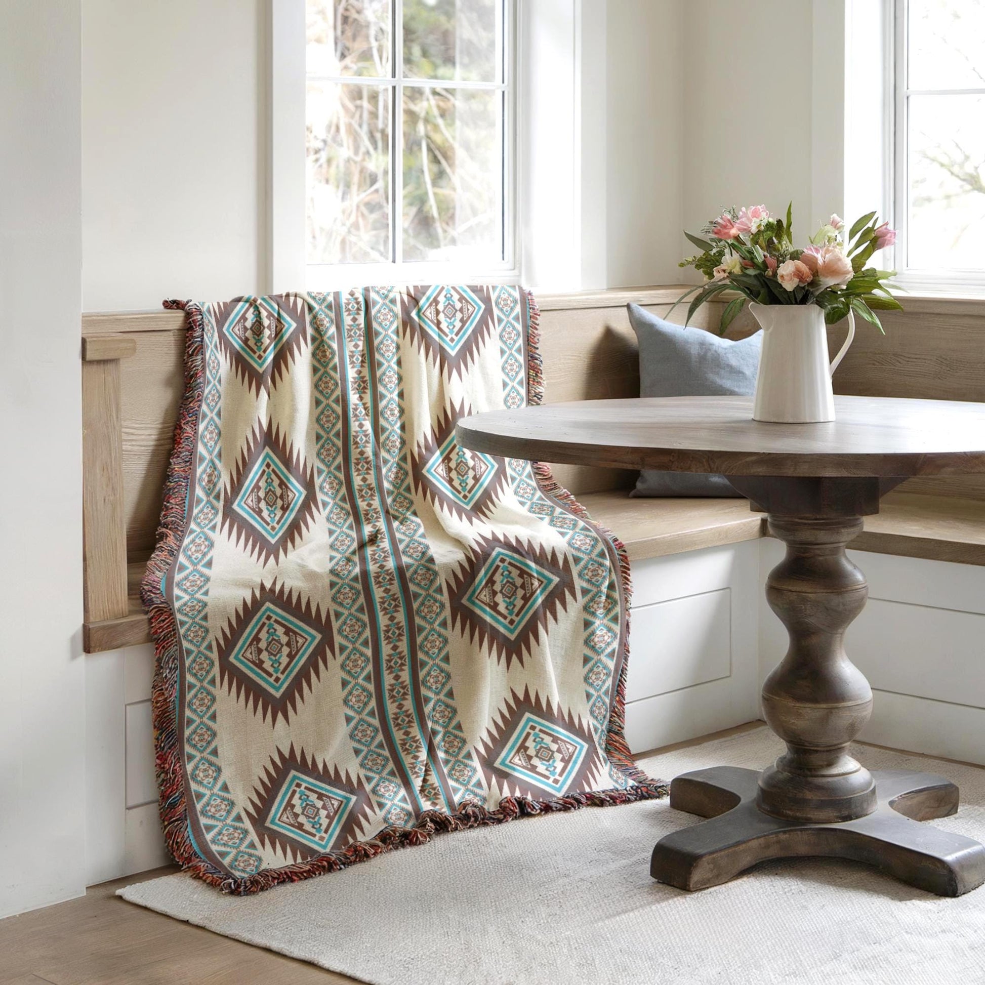 a cozy corner with a wooden table, a bench, and a colorful throw blanket draped over the bench. A vase of flowers is placed on the table, adding a touch of nature to the space. The room has a clean, minimalist aesthetic with white walls and a wooden floor.