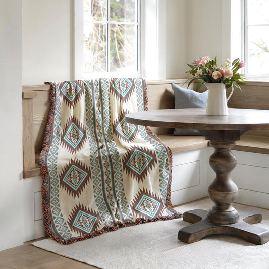 a cozy corner with a wooden table, a bench, and a colorful throw blanket draped over the bench. A vase of flowers is placed on the table, adding a touch of nature to the space. The room has a clean, minimalist aesthetic with white walls and a wooden floor.