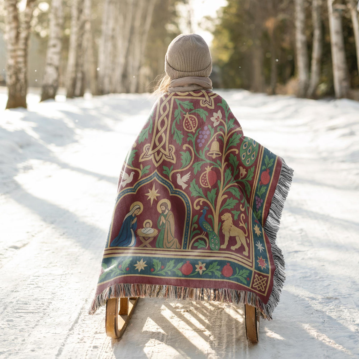 A person is sitting on a sled in a snowy forest, wearing a colorful blanket with a religious design.