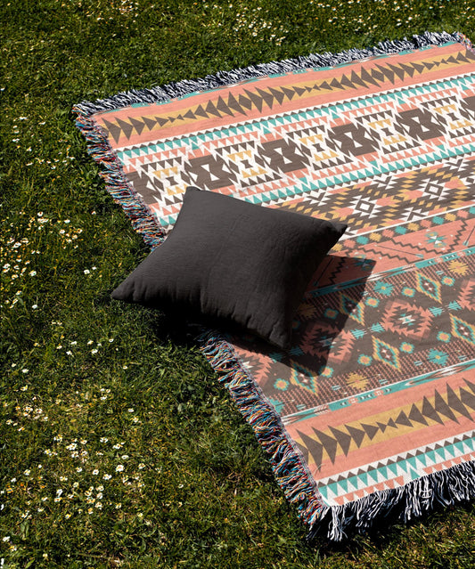 a colorful, patterned blanket with fringed edges and a single black pillow resting on it, placed on a grassy lawn with white flowers.
