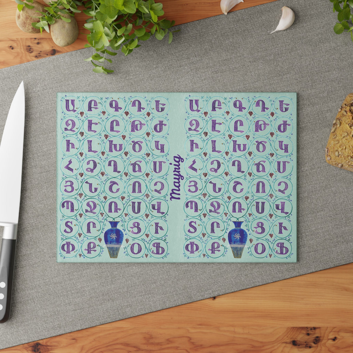 Mint Armenian alphabet cutting board with blue vases on a grey mat beside a knife and fresh herbs