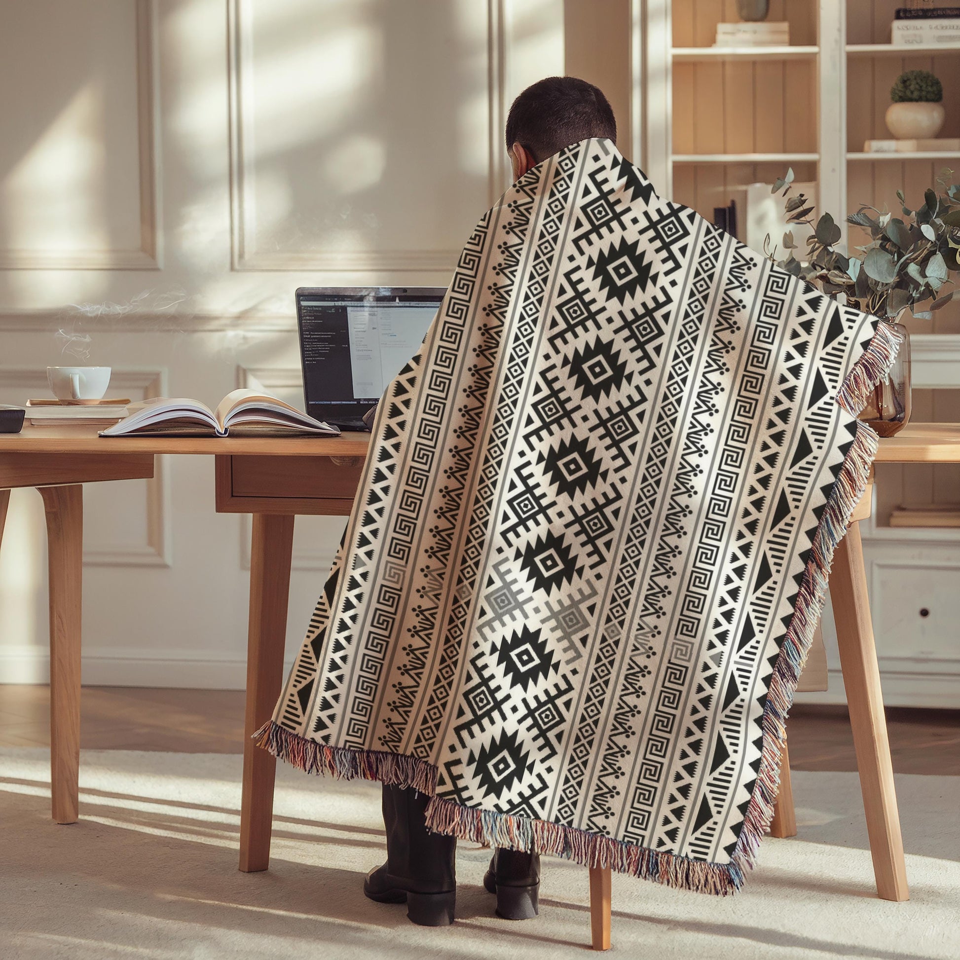 A person is sitting at a desk, wrapped in a patterned blanket, using a laptop computer.