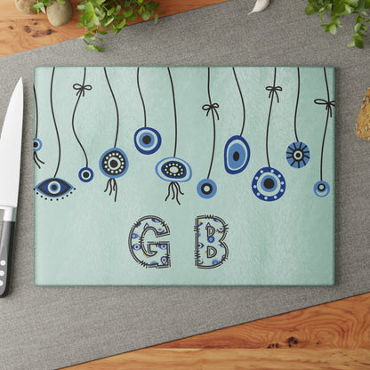 Mint evil eye cutting board with hanging beads on a grey mat beside a knife and fresh herbs