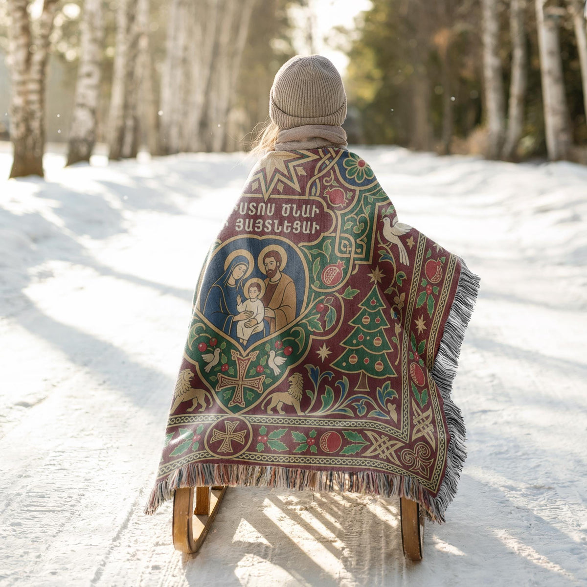 A person is sitting on a sled in a snowy forest, wrapped in a colorful blanket with a religious design.