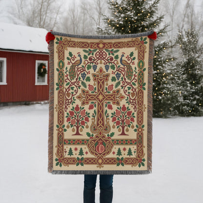 A person is holding up a large, ornate, and colorful blanket with a cross design in front of a snowy landscape with a red building and Christmas trees in the background.