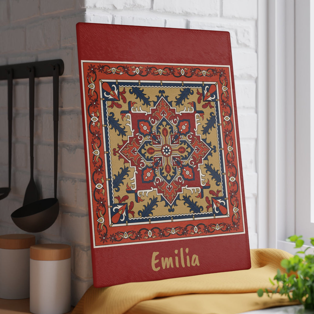 Red Armenian rug cutting board with a gold name on a kitchen surface with herbs and stones nearby