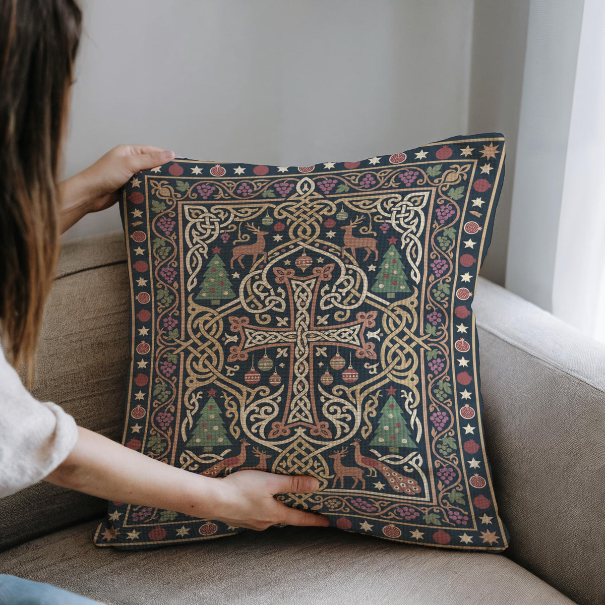 A person is holding up a decorative pillow with an intricate, colorful design featuring a cross, trees, and other festive elements.